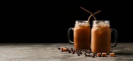 Mason jars of tasty iced coffee with milk, beans and sugar cubes on dark background with space for...