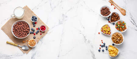 Different breakfast cereals, milk and berries on light marble background with space for text