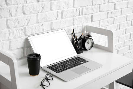 Modern laptop and paper cup of coffee on table near white brick wall