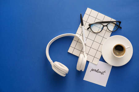 Headphones, eyeglasses, notebook, cup of coffee and paper sheet with word PODCAST on blue backgroundの写真素材