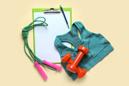 Clipboard with pen, sport top, dumbbells and skipping rope on beige background