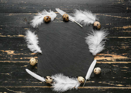 Composition with beautiful white feathers, quail eggs and board on black wooden background