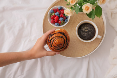 Woman taking bun from tray with breakfast on bed, closeup
