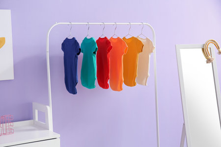 Rack with colorful baby bodysuits near lilac wall in children's room