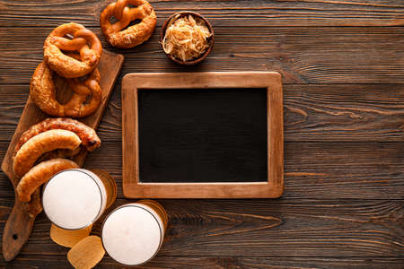 Glasses of cold beer, blank chalkboard, board with Bavarian sausages and snacks on wooden...