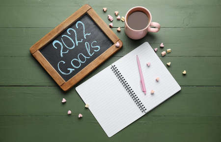 Board with text 2022 GOALS, notebook, candies and cup of coffee on green wooden background
