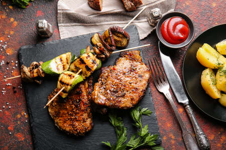 Board with tasty pork steaks and grilled vegetables on grunge background
