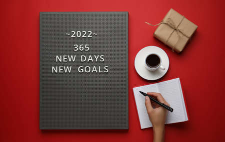 Board with text 365 NEW DAYS NEW GOALS and female hand with notebook and pen on color background
