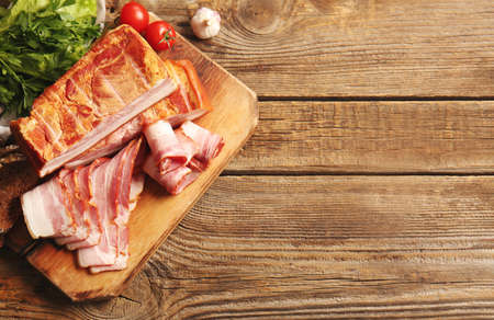 Board with tasty smoked bacon and fresh vegetables on wooden background