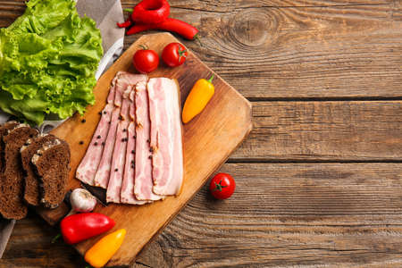 Board with slices of tasty smoked bacon and fresh vegetables on wooden background