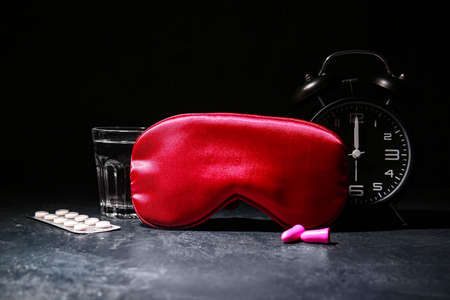 Sleep mask, alarm clock, pills and earplugs on dark background