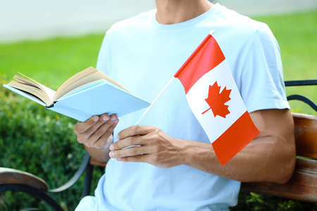 Male student with flag of Canada reading book while sitting on bench outdoorsの写真素材