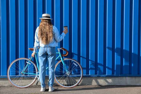 Pretty teenage girl with cup of coffee and bicycle near blue fence