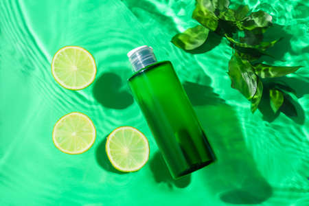 Bottle of cosmetic product, lime slices and plant branch in water on color background