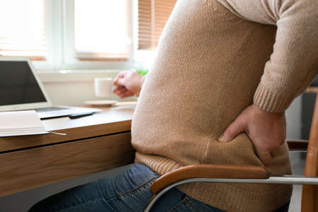Senior man with cup of coffee suffering from back pain in chair at homeの写真素材