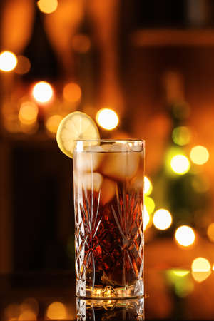 Glass of tasty Cuba Libre cocktail on table against blurred lightsの写真素材