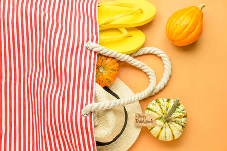 Beach bag with flip flops, wicker hat, pumpkins and paper card with text HAPPY THANKSGIVING on color backgroundの写真素材