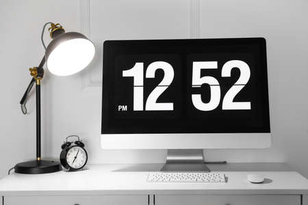 Modern computer, alarm clock and glowing lamp on desk in officeの写真素材