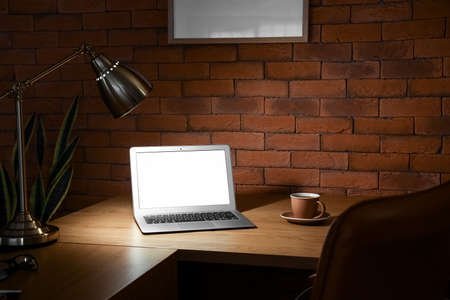 Modern workplace with laptop, cup of coffee and glowing lamp near brick wall in dark officeの写真素材