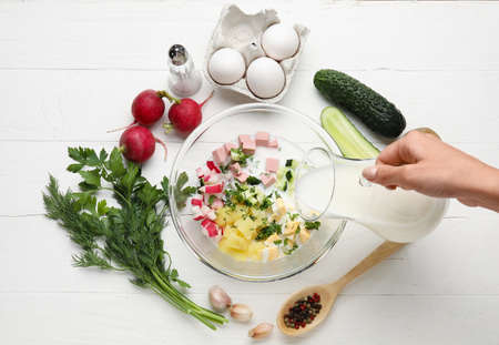 Woman pouring kefir into bowl with ingredients for okroshka on white wooden backgroundの写真素材