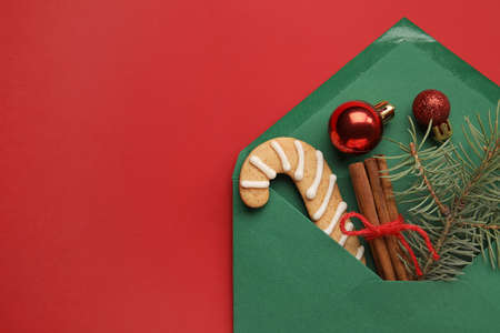 Envelope with fir tree branch, cinnamon, balls and cookies on red background, closeup