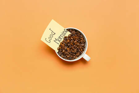 Cup with coffee beans and sticky note with text GOOD MORNING on color backgroundの写真素材