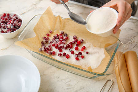 Woman sprinkling sugar onto cranberry on light background, closeup