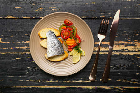 Plate of tasty sea bass fish with vegetables on black wooden background