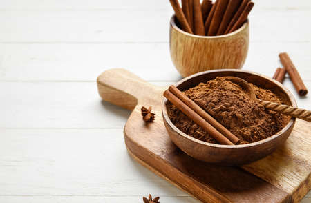 Bowl with cinnamon powder and sticks on white wooden background