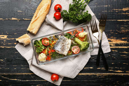 Baking dish of tasty sea bass fish with vegetables on black wooden background