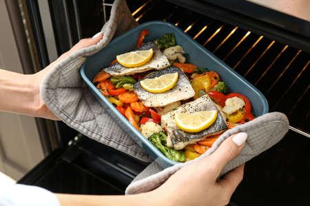 Woman taking out from oven baking dish with tasty sea bass fish in kitchenの写真素材