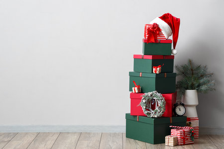 Stack of Christmas gift boxes, alarm clock and vase with fir tree branches on floor near white wall
