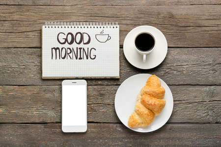 Notebook with text GOOD MORNING, mobile phone, cup of coffee and croissant on wooden background