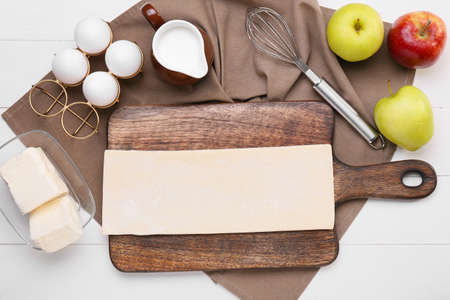 Board with fresh dough and ingredients for apple strudel on white wooden background