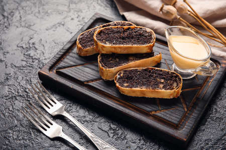 Wooden board with pieces of tasty poppy seed strudel on black background
