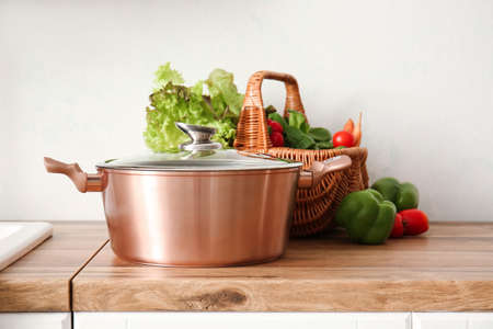 Shiny cooking pot and basket with fresh vegetables on counter near light wallの写真素材