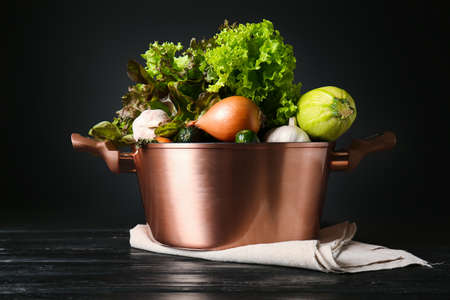 Shiny cooking pot with fresh vegetables on wooden table against dark backgroundの写真素材