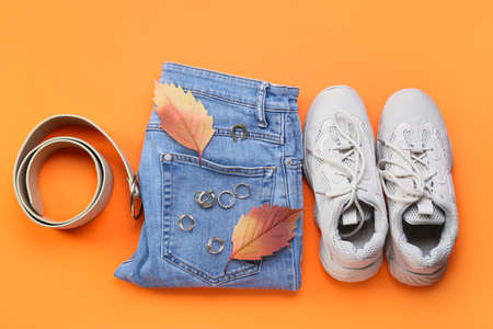 Belt, jeans, shoes, jewelry and dry leaves on color background