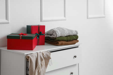 Chest of drawers with Christmas gift boxes and warm clothes near light wall