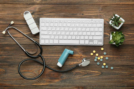 Asthma inhaler, keyboard, stethoscope and pills on wooden background