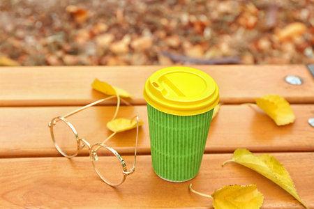 Takeaway cup of tasty coffee and eyeglasses on bench in autumn parkの写真素材