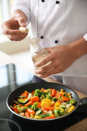 Male chef with bottle of oil frying vegetables in kitchen, closeup