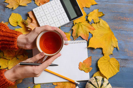Woman with cup of tea, notebook and autumn decor on blue wooden background