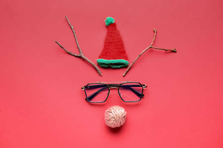 Creative composition with reindeer face made of Christmas hat, glasses, branches and rope on red backgroundの写真素材