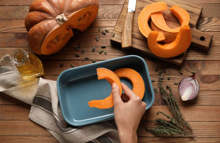 Woman putting fresh pumpkin pieces into baking dish on wooden backgroundの写真素材