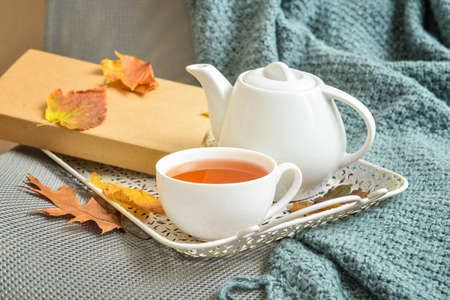 Tray with tea pot, cup of hot beverage and autumn leaves on sofa in roomの写真素材