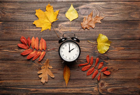 Alarm clock and autumn leaves on wooden background. daylight saving time end