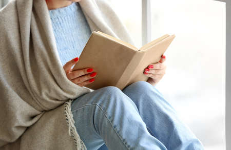 Beautiful woman in plaid reading book on windowsill at home, closeup