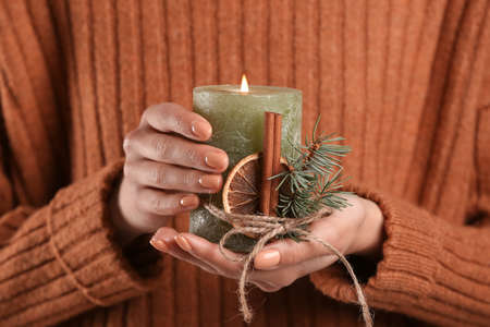 Woman in warm sweater holding burning candle with fir branches, dried citrus slice and cinnamon, closeupの写真素材