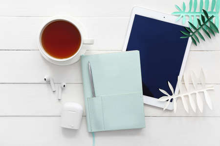 Stylish notebook with tablet computer, earphones, cup of tea and paper palm leaves on white wooden...